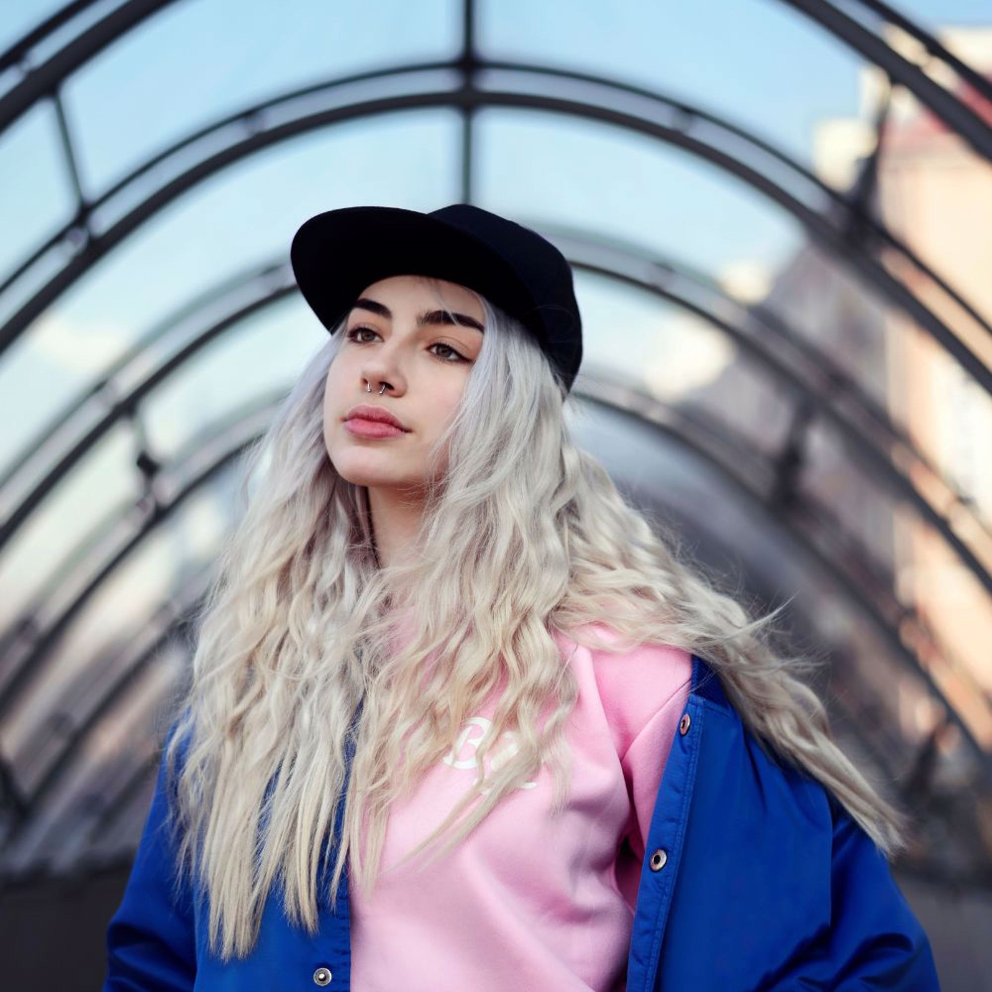 A young woman with long, wavy, cool blonde hair stands confidently in an archway with a modern glass design. She is wearing a black baseball cap, a blue jacket, and a pink shirt, looking upward with a calm expression. Her stunning hair color is thanks to #10.21 Pearl Blonde Permanent Hair Dye by Oiamiga.